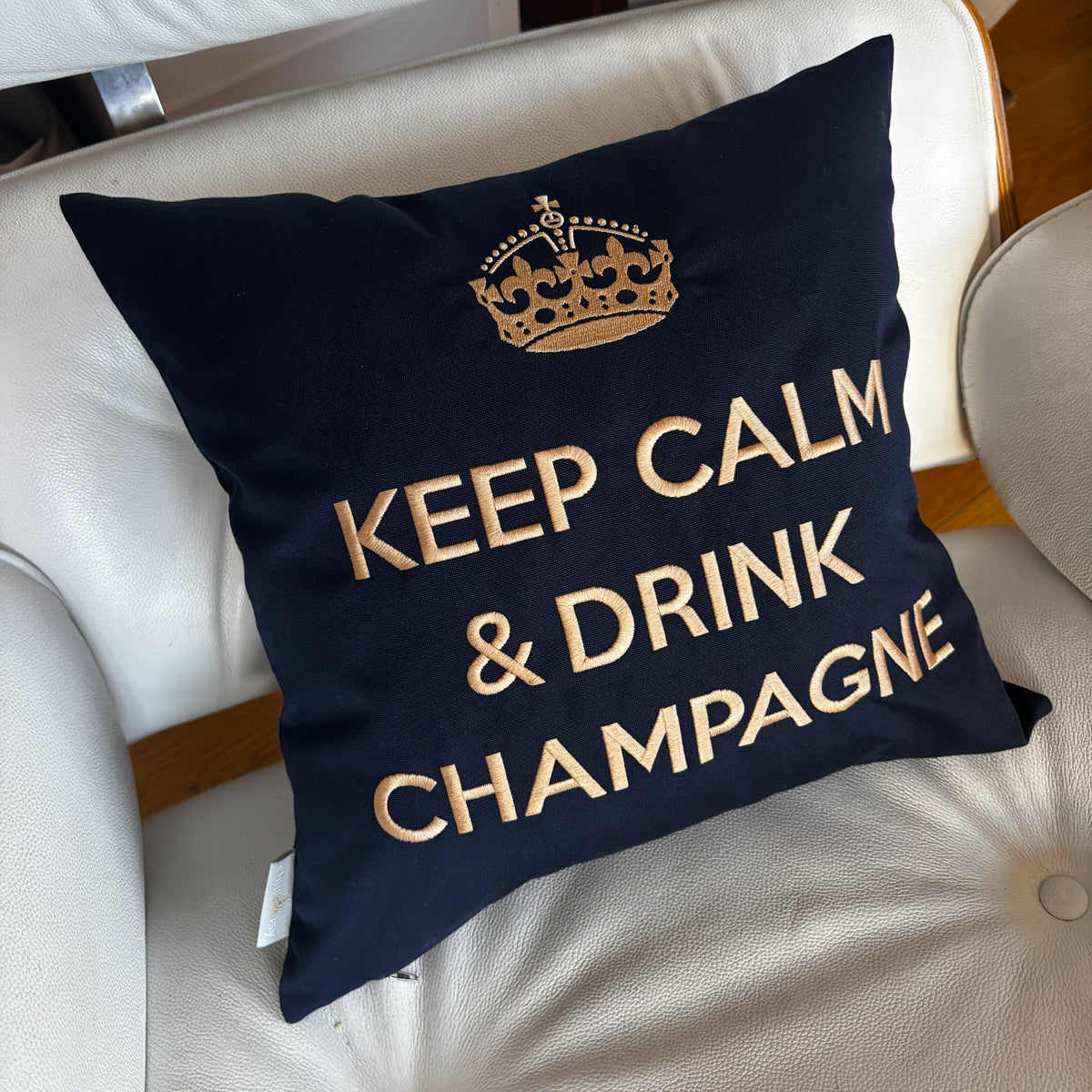 Dunkelblaues Dekokissen mit beiger Stickerei: eine Krone über dem Schriftzug "KEEP CALM & DRINK CHAMPAGNE", liegend auf einem hellen VITRA Ledersessel.