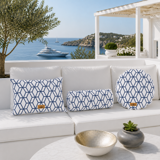 Exklusive Outdoor-Kissen-Kollektion von chillisy® auf einer weißen Luxus-Gartenlounge mit Meerblick. Rechteckiges Dekokissen, Nackenrolle und rundes Kissen mit weißem Stoff, marineblauem geometrischem Muster, weißem Keder und goldenem chillisy®-Label – stilvoll inszeniert vor einer mediterranen Küstenlandschaft mit Yacht.