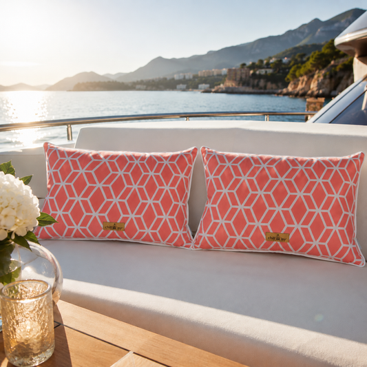 Zwei luxuriöse chillisy® Outdoor-Kissen mit korallfarbenem geometrischem Muster, weißem Keder und goldenem chillisy®-Label auf einer eleganten weißen Yacht-Lounge mit Blick auf das Meer bei warmem Abendlicht.