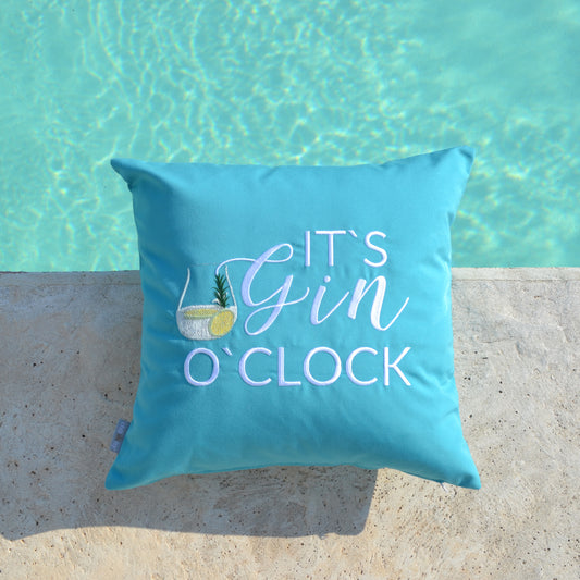 Türkises Kissen mit weißem Stick IT'S GIN O'CLOCK und Gin Tonic Glas auf Terrassenboden am Pool liegend.