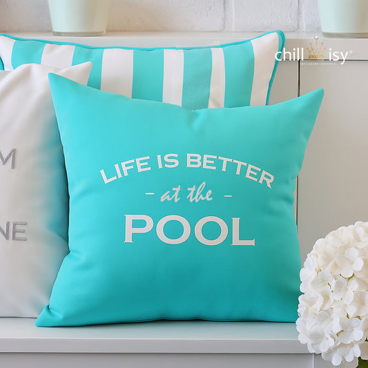 Dekokissen in Türkis mit weißer Stickerei LIFE IS BETTER at the POOL auf einer Bank mit weißem Hintergrund und anderen Kissen abgebildet.