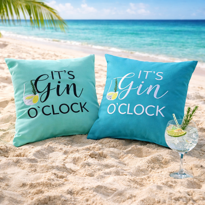 Zwei Dekokissen in der Farbe Mint und Türkis jeweils mit der Aufschrift „IT‘S GIN O‘CLOCK“ und Gin Glas am Sandstrand mit Meer.