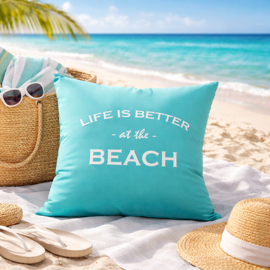 Türkises Dekokissen mit weißem Stick „LIFE IS BETTER at the BEACH“ am Strand mit Sonnenhut, Badetasche, Flip Floos und Palme.
