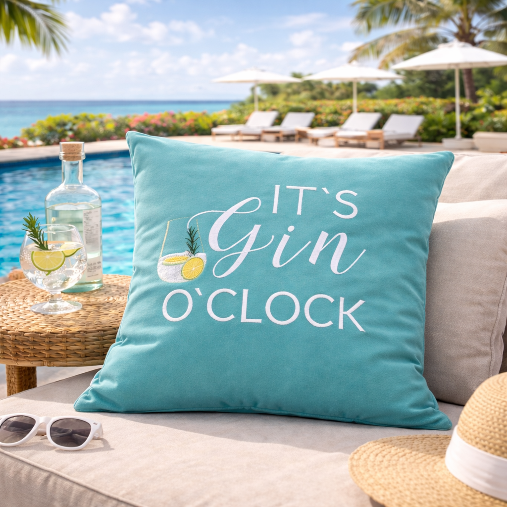 Türkises Kissen mit weißem Stick IT'S GIN O'CLOCK und Gin Tonic Glas auf Sofa am Pool liegend.