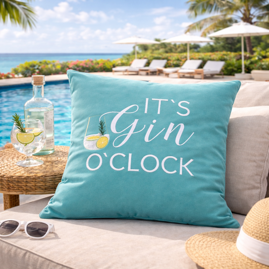 Türkises Kissen mit weißem Stick IT'S GIN O'CLOCK und Gin Tonic Glas auf Sofa am Pool liegend.
