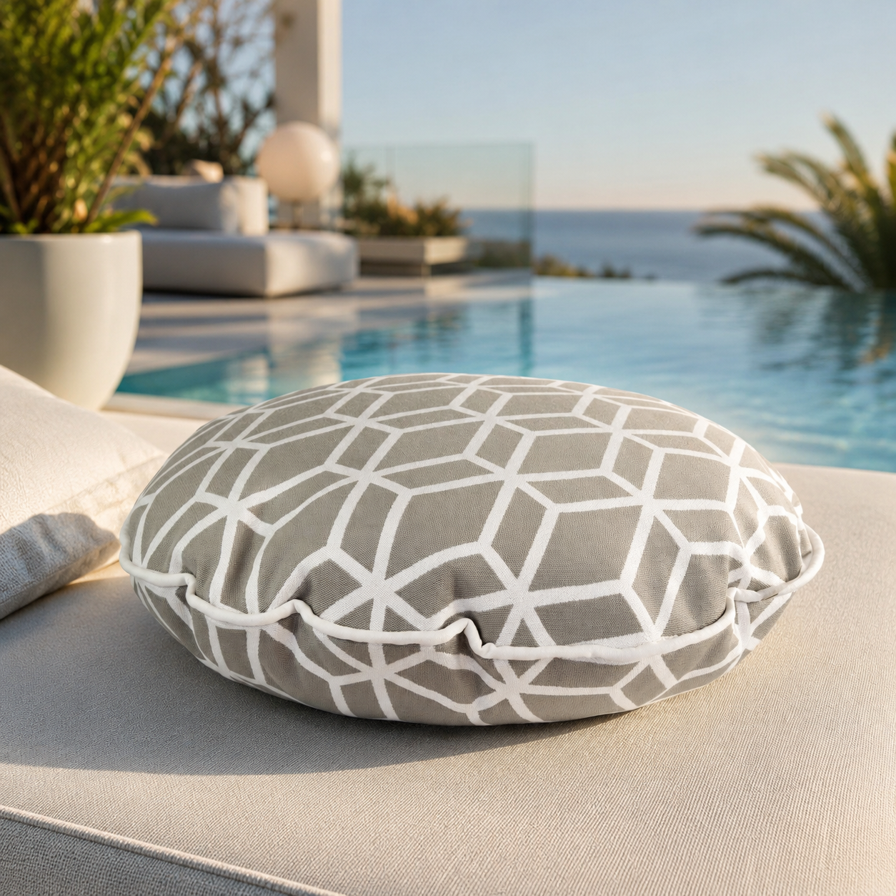 chillisy® Designer-Rundkissen Crystal Gold Hellgrau mit weißem geometrischem Muster, weißem Keder und Goldlabel auf einer luxuriösen Outdoor-Lounge mit Infinity-Pool und Meerblick – Premium Outdoor-Kissen, genäht in Deutschland