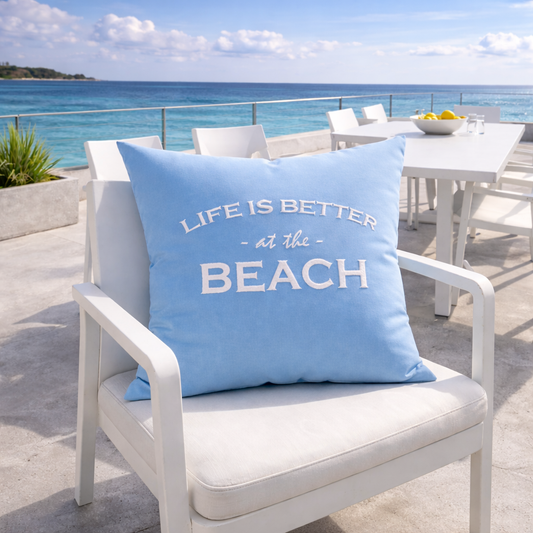 Dekokissen Aqua Blau mit weißer Stickerei LIFE IS BETTER AT THE BEACH auf Terrasse mit Meerblick auf weißem Stuhl