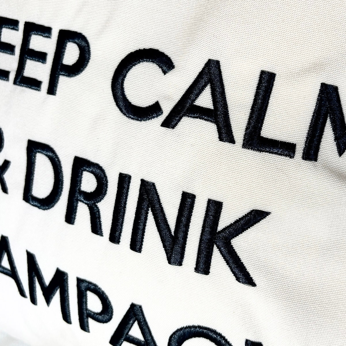 Quadratisches, cremefarbenes Dekokissen mit einem schwarzen Kronensymbol über der aufgestickten Aufschrift „KEEP CALM & DRINK CHAMPAGNE“.