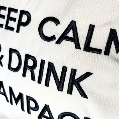 Quadratisches, cremefarbenes Dekokissen mit einem schwarzen Kronensymbol über der aufgestickten Aufschrift „KEEP CALM & DRINK CHAMPAGNE“.