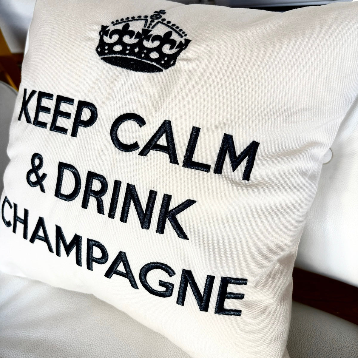 Quadratisches, cremefarbenes Dekokissen mit einem schwarzen Kronensymbol über der aufgestickten Aufschrift „KEEP CALM & DRINK CHAMPAGNE“.