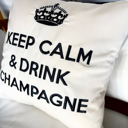 Quadratisches, cremefarbenes Dekokissen mit einem schwarzen Kronensymbol über der aufgestickten Aufschrift „KEEP CALM & DRINK CHAMPAGNE“.