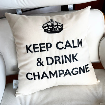 Quadratisches, cremefarbenes Dekokissen mit einem schwarzen Kronensymbol über der aufgestickten Aufschrift „KEEP CALM & DRINK CHAMPAGNE“ auf weißem Vitra Sessel.