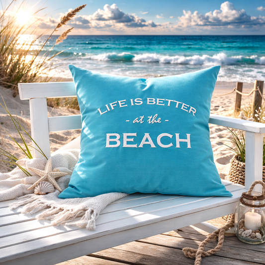 Türkises Dekokissen am Strand mit weißem Stick „LIFE IS BETTER at the BEACH“ am Strand auf einer weißen Bank mit Muscheln und Kerze,