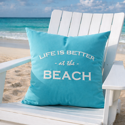 Türkises Dekokissen mit weißem Stick „LIFE IS BETTER at the BEACH“ am Strand auf weißem Strandsessel.