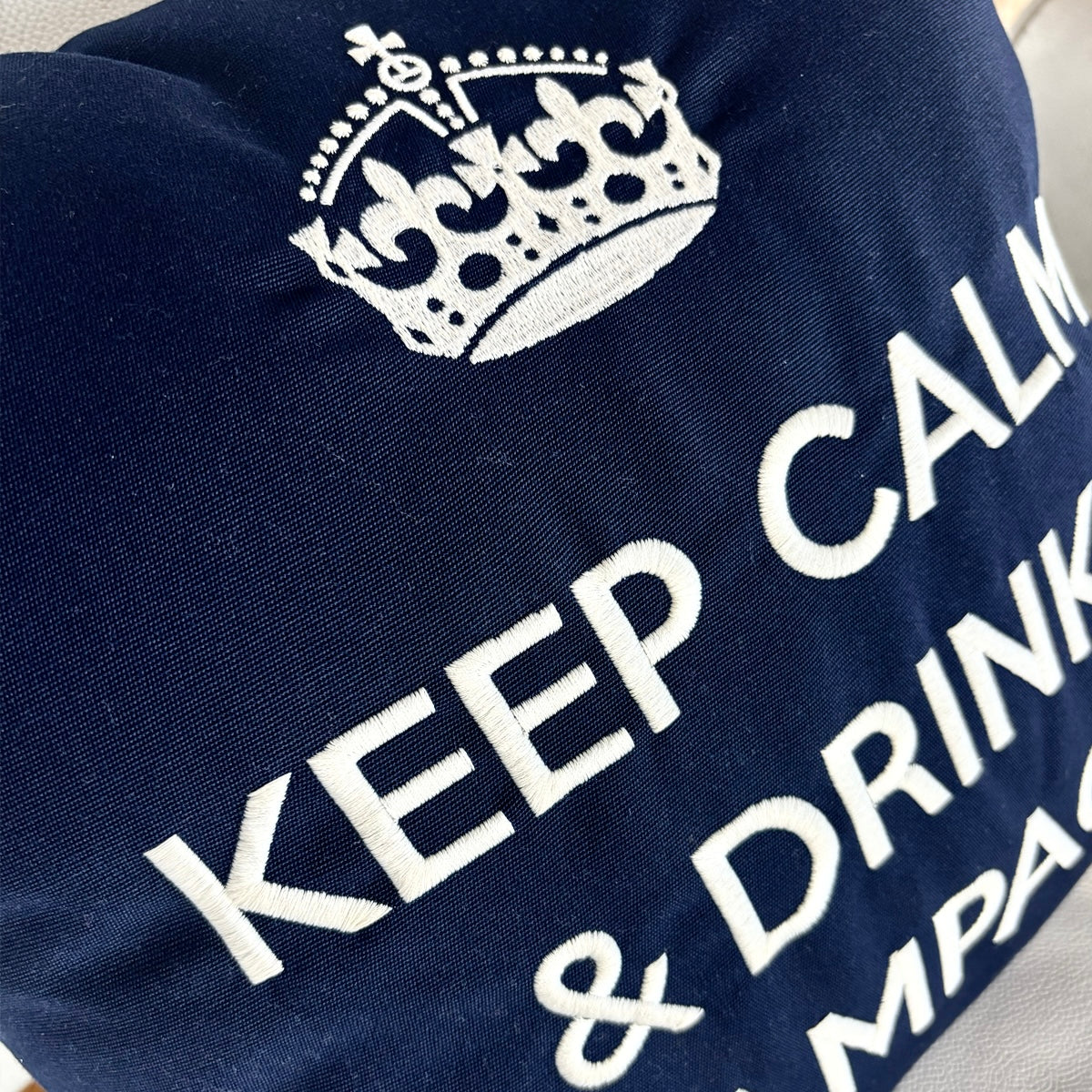 Dunkelblaues Dekokissen mit weißer Stickerei einer Krone und dem Schriftzug „KEEP CALM & DRINK CHAMPAGNE“, liegend auf einem hellen Ledersessel.