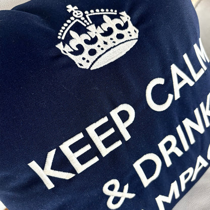 Dunkelblaues Dekokissen mit weißer Stickerei einer Krone und dem Schriftzug „KEEP CALM & DRINK CHAMPAGNE“, liegend auf einem hellen Ledersessel.