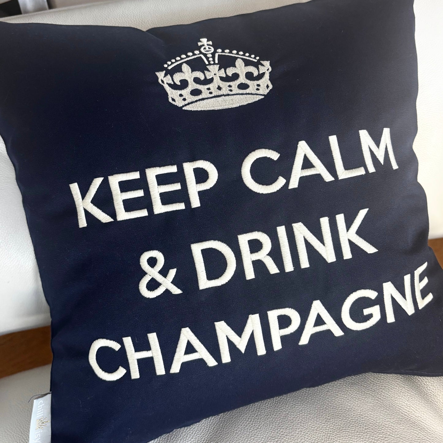 Dunkelblaues Dekokissen mit weißer Stickerei einer Krone und dem Schriftzug „KEEP CALM & DRINK CHAMPAGNE“, liegend auf hellen VITRA Ledersessel.