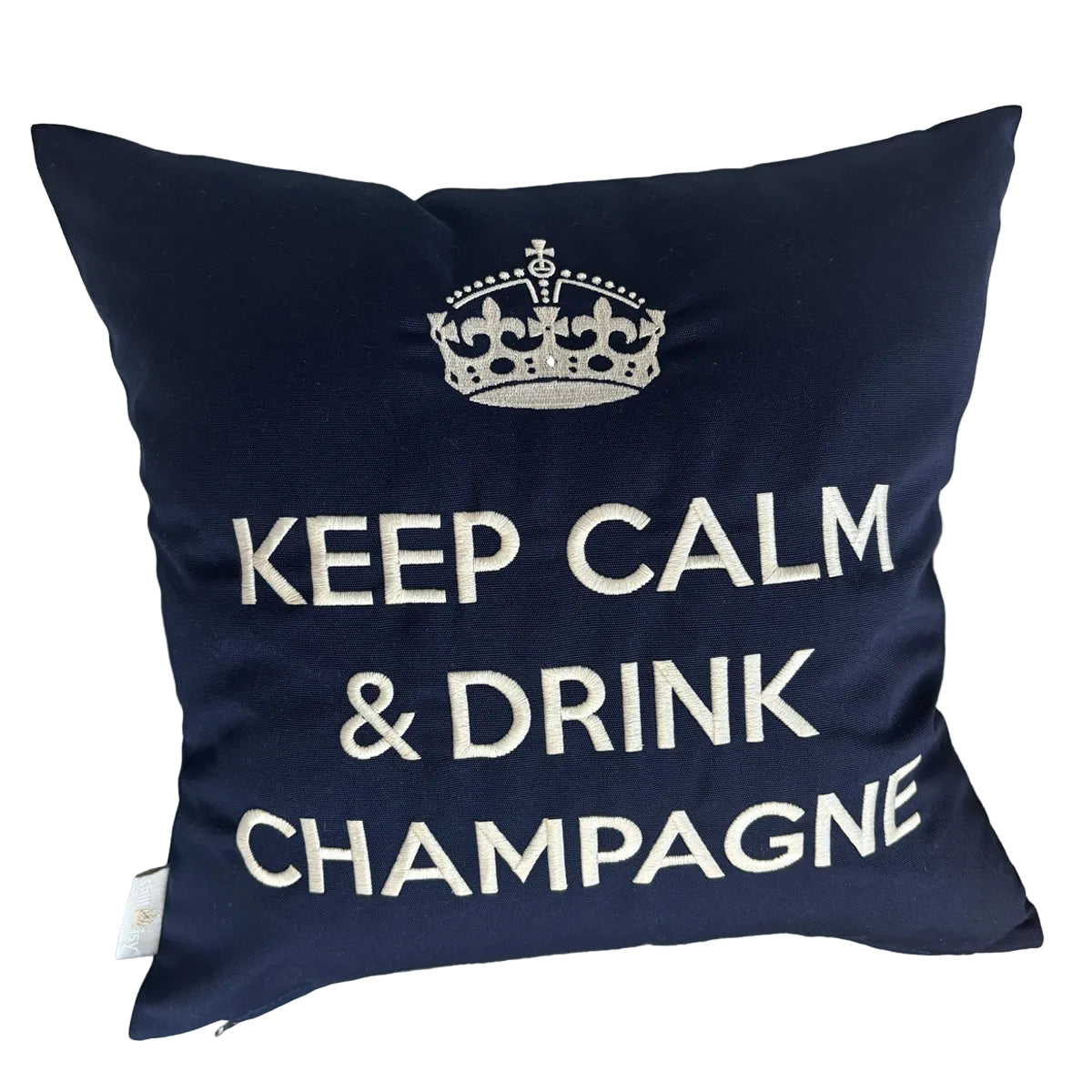 Dunkelblaues Dekokissen mit weißer Stickerei einer Krone und dem Schriftzug „KEEP CALM & DRINK CHAMPAGNE“, liegend auf einem hellen Ledersessel.