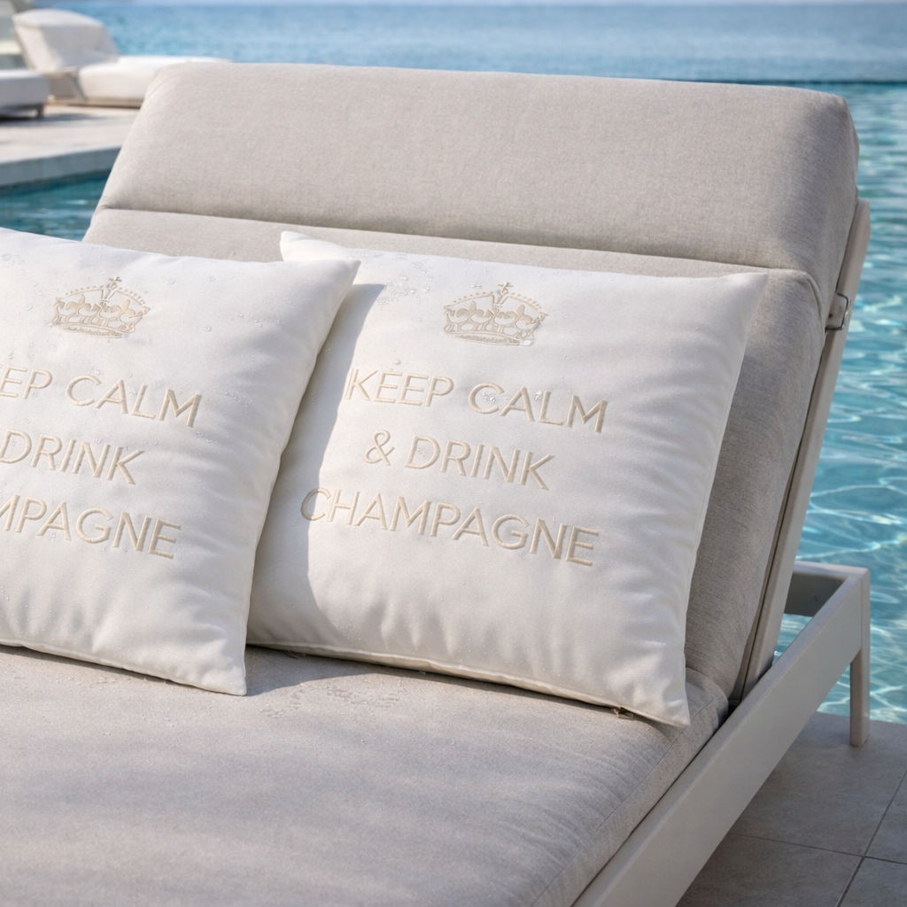 Weiße Outdoor-Dekokissen mit Wollweste Stickerei „Keep Calm & Drink Champagne“ auf moderner hellgrauer Sonnenliege am Pool in luxuriöser Umgebung.