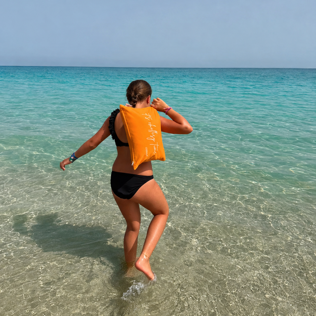 Frau trägt das orange Piccolino Schwimm- und Reisekissen von chillisy® auf dem Weg ins Meer