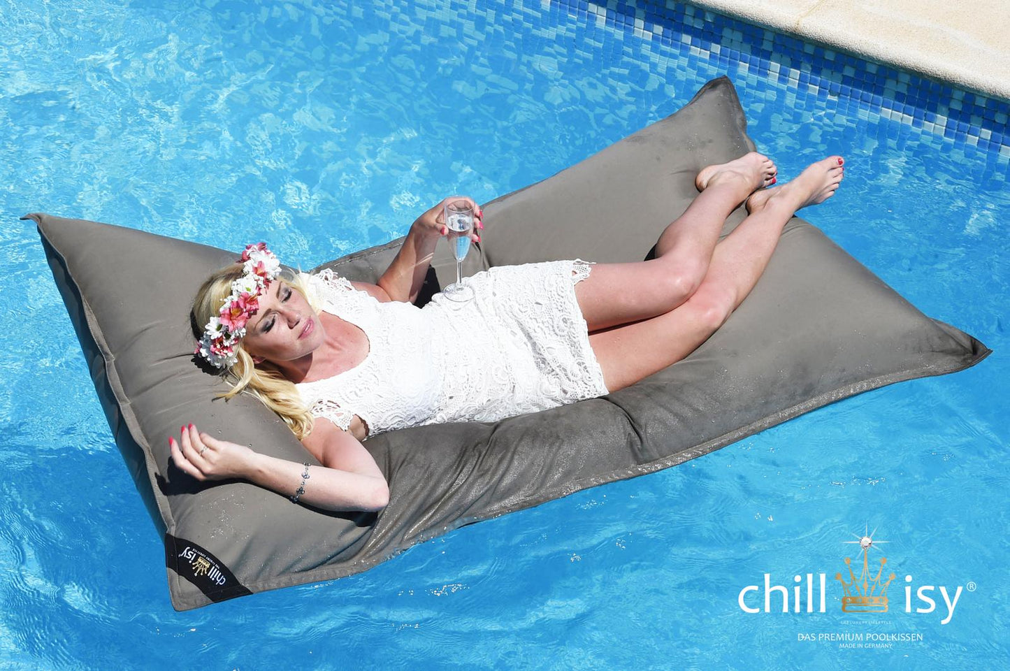Taupe farbener Pool Sitzsack Mallorca in der Größe Lungo 190x100 cm von chillisy schwimmend auf einem Swimmingpool – mit Frau darauf im weißen Kleid und Blumenkranz auf dem Kopf. Hergestellt in Deutschland.